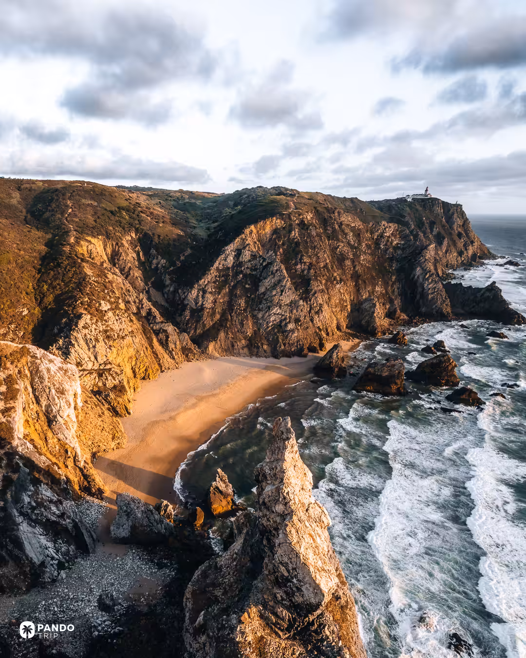 Praia da Ursa beach in shadow below sunlit cliffs, with the Cabo da Roca lighthouse in the distance