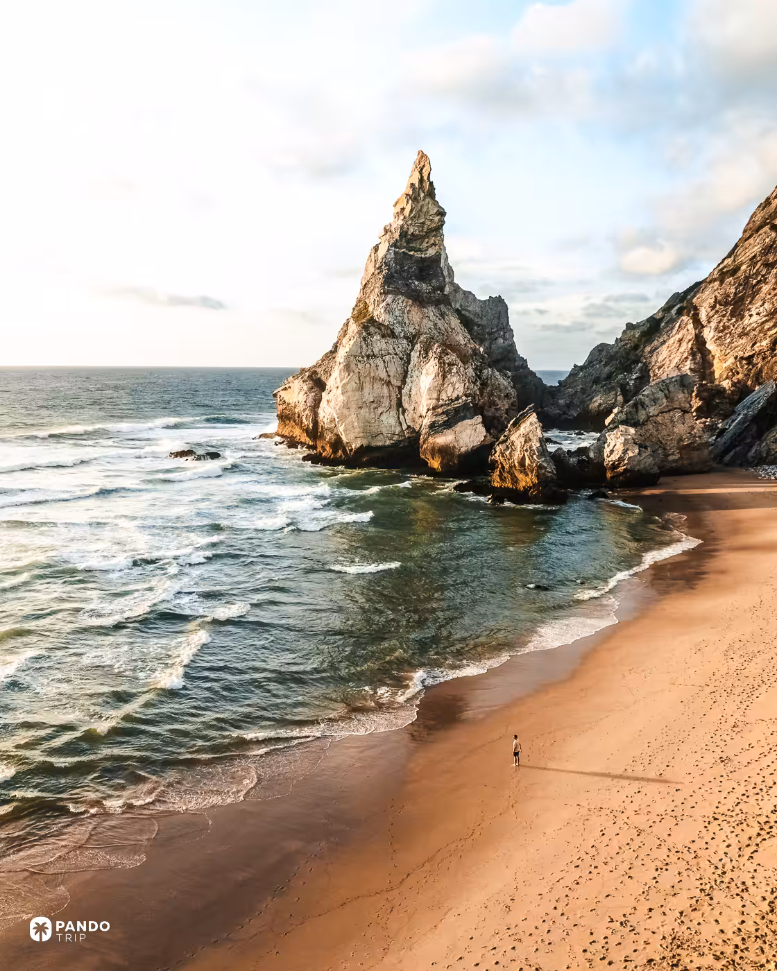 Sharp limestone spire and calm green water at Praia da Ursa with a single person on the beach