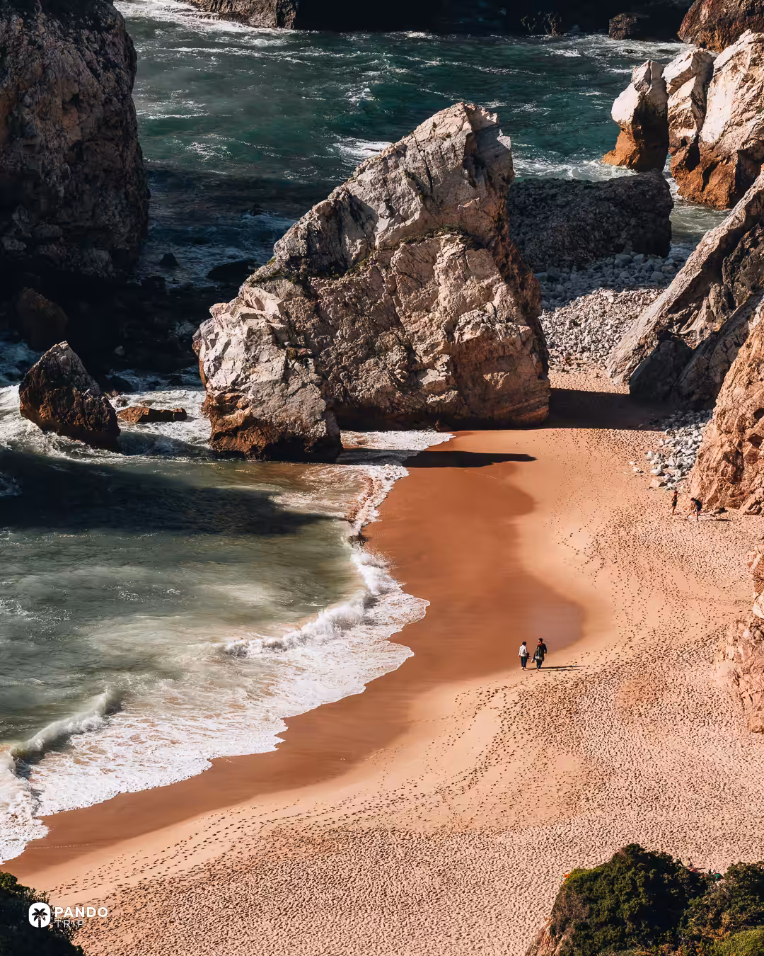 A couple walking along the curved shoreline of Praia da Ursa below a large boulder