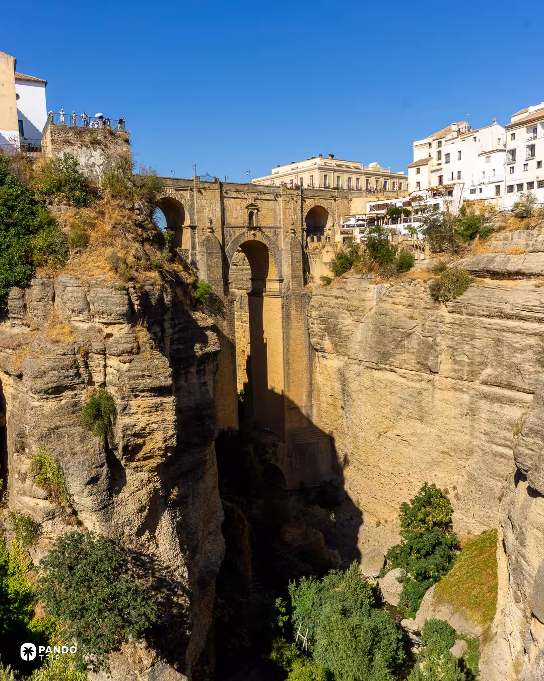 Looking down into the El Tajo gorge in Ronda towards the arches of the Puente Nuevo, with visitors on the viewpoint above.