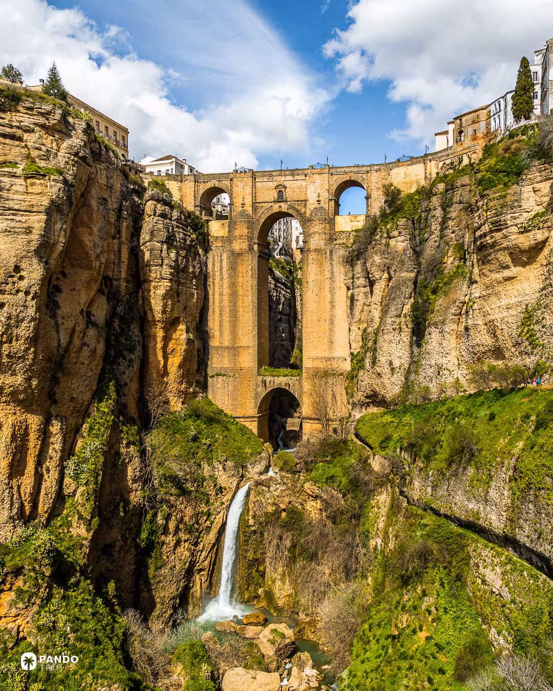 The Puente Nuevo in Ronda seen from below, its tall stone piers rising from the green floor of the El Tajo gorge above a waterfall.