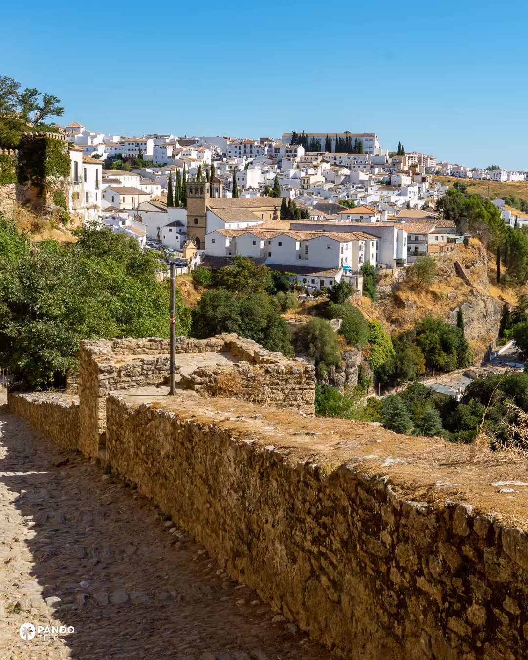 Old stone walls and a cobbled path on the edge of Ronda, with the white town and a church tower beyond.