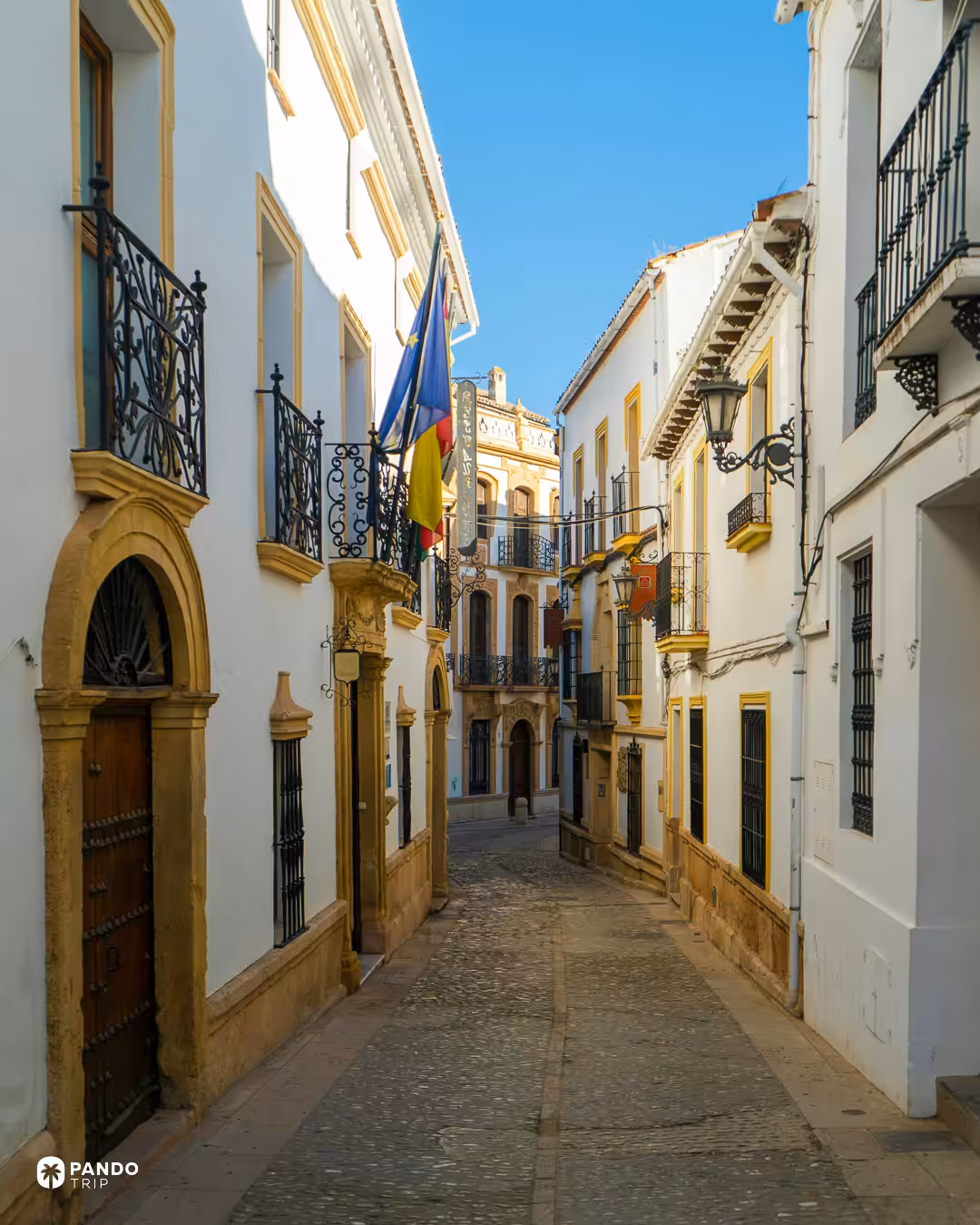 A cobbled street in Ronda with ornate doorways, iron balconies and flags on a building facade.