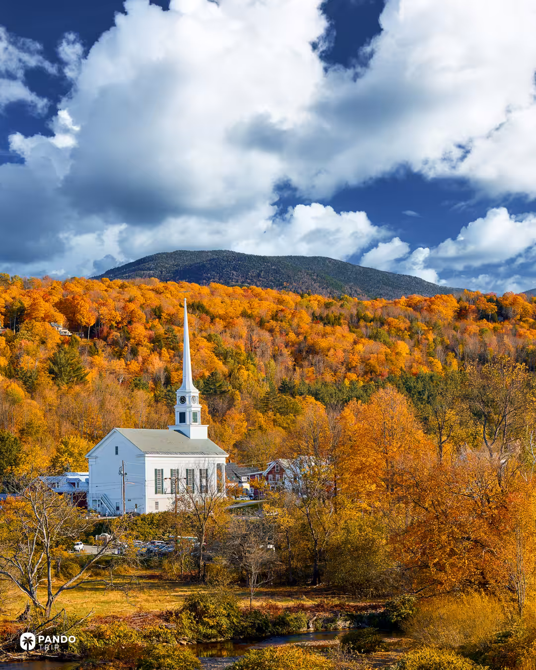 The white Stowe Community Church and its tall spire in front of a hillside of orange autumn trees and a mountain under dramatic clouds.